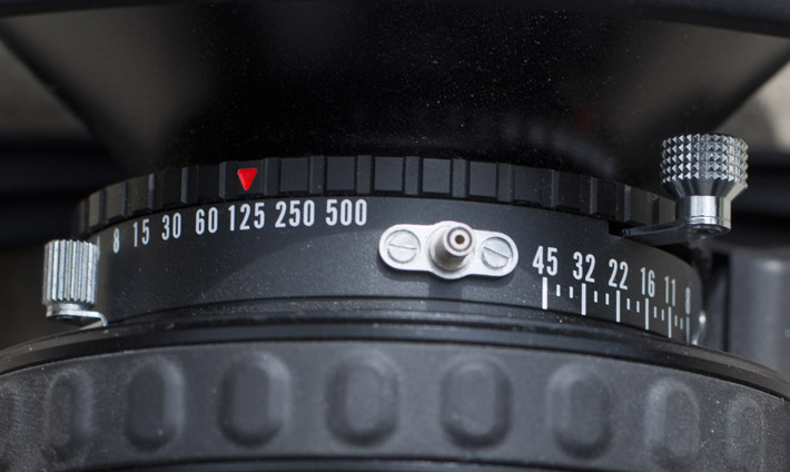 Use the knob on the left to adjust the aperture (the needle barely visible on the right). The black ring with the red arrow is used to select shutter speed. And the right knob cocks the shutter when pulled all the way down.